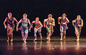 Twyla Tharp dancers in a line stepping forward in colorful costumes