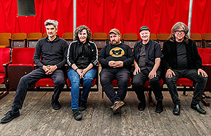 Magnetic Fields band members sitting in a line in red auditorium seats. All members are wearing black shirts and looking at the camera