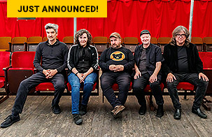 Magnetic Fields band members sitting in a line in red auditorium seats. All members are wearing black shirts and looking at the camera